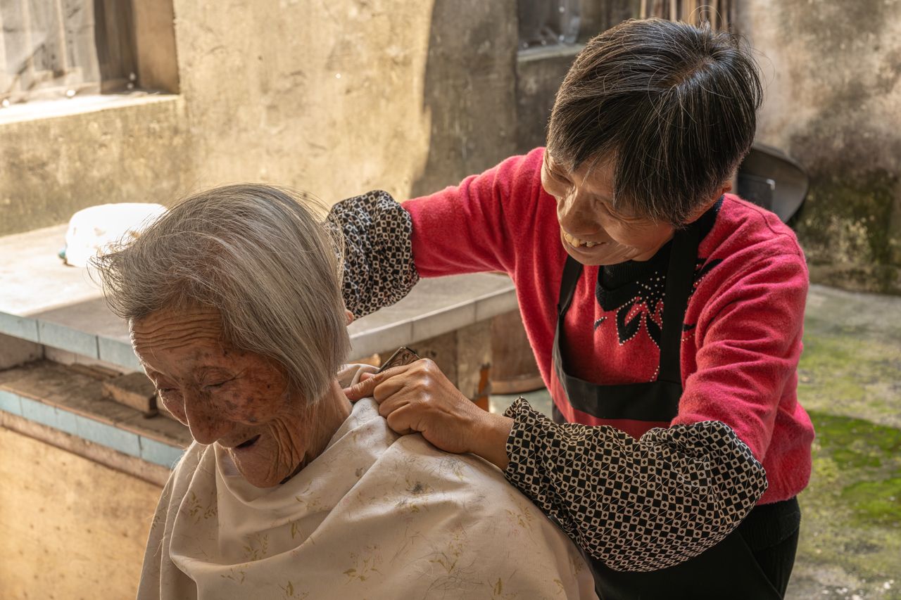 services-04 An elderly woman receives a haircut from another woman in a rustic outdoor setting.